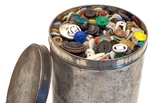 A close-up view of a round, silver metal trash can filled to the top with a variety of plastic and metal buttons of different shapes, sizes, and colours, including blue, black, white, green, and brown. The trash can is positioned outdoors on a paved surface with natural daylight illuminating the objects inside. The can's lid is resting against its side, partially visible on the left. This scene relates to waste collection and rubbish disposal services in Raynes Park, highlighting the types of small household items managed during garden clearance or rubbish removal operations by Garden Clearance Raynes Park, with the background suggesting a typical urban environment in South West London.