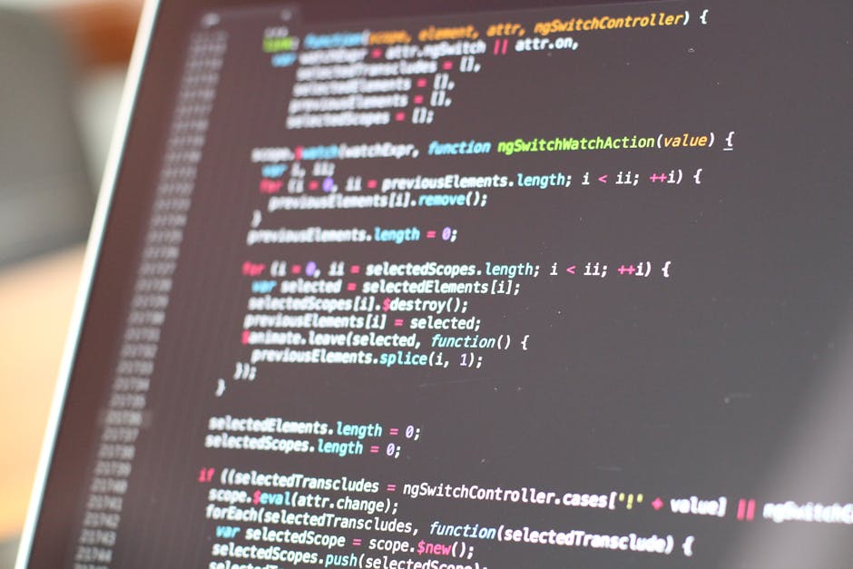 A close-up view of a person working on a laptop placed on a dark desk or table. The laptop screen displays a code editor with lines of programming code in various colors, including green, blue, and wh