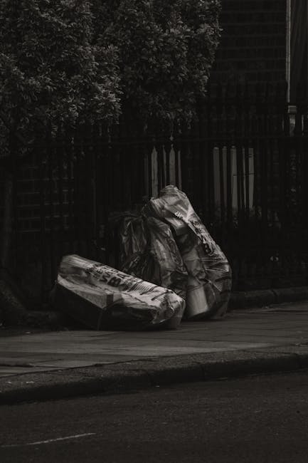 Three large black plastic rubbish bags, partly torn and crumpled, are stacked on a curb alongside a sidewalk in an outdoor setting. The bags appear filled with waste or disposed items and are position