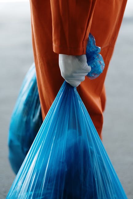 Three large black plastic rubbish bags, partly torn and crumpled, are stacked on a curb alongside a sidewalk in an outdoor setting. The bags appear filled with waste or disposed items and are position
